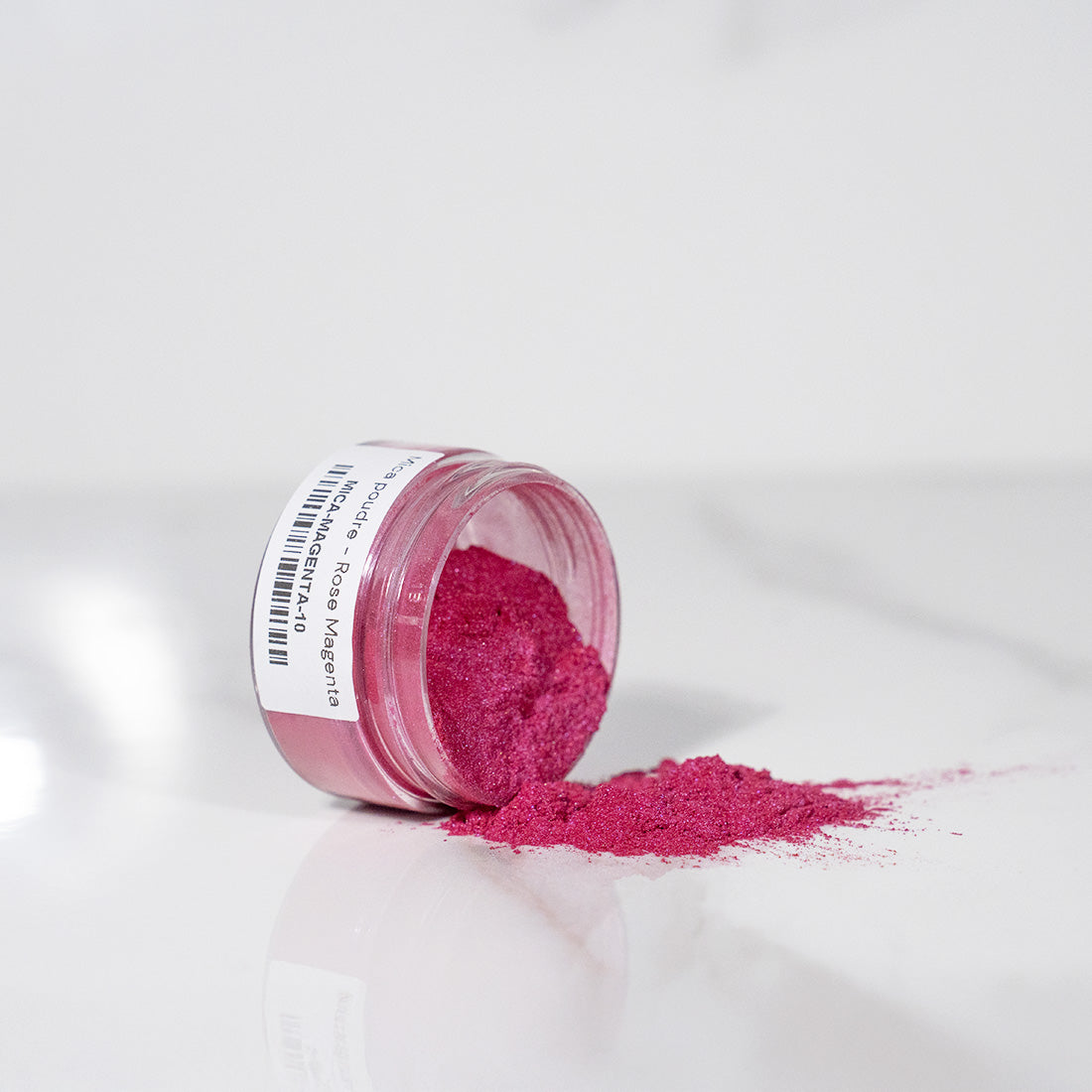 Pot transparent renversé avec de la poudre de mica rose magenta scintillant sur un plan blanc