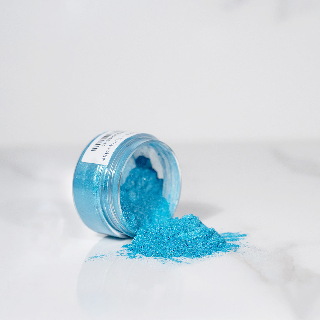 Pot transparent renversé avec de la poudre de mica turquoise scintillant sur un plan blanc