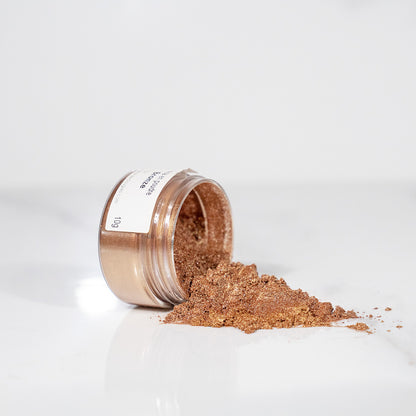 Pot transparent renversé avec de la poudre de mica bronze scintillant sur un plan blanc