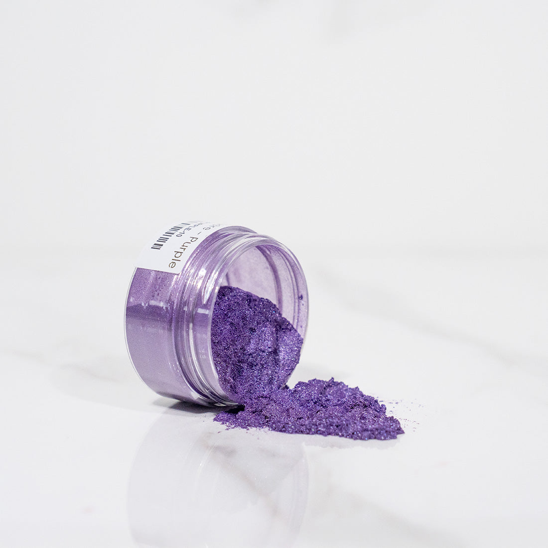 Pot transparent renversé avec de la poudre de mica violet scintillant sur un plan blanc
